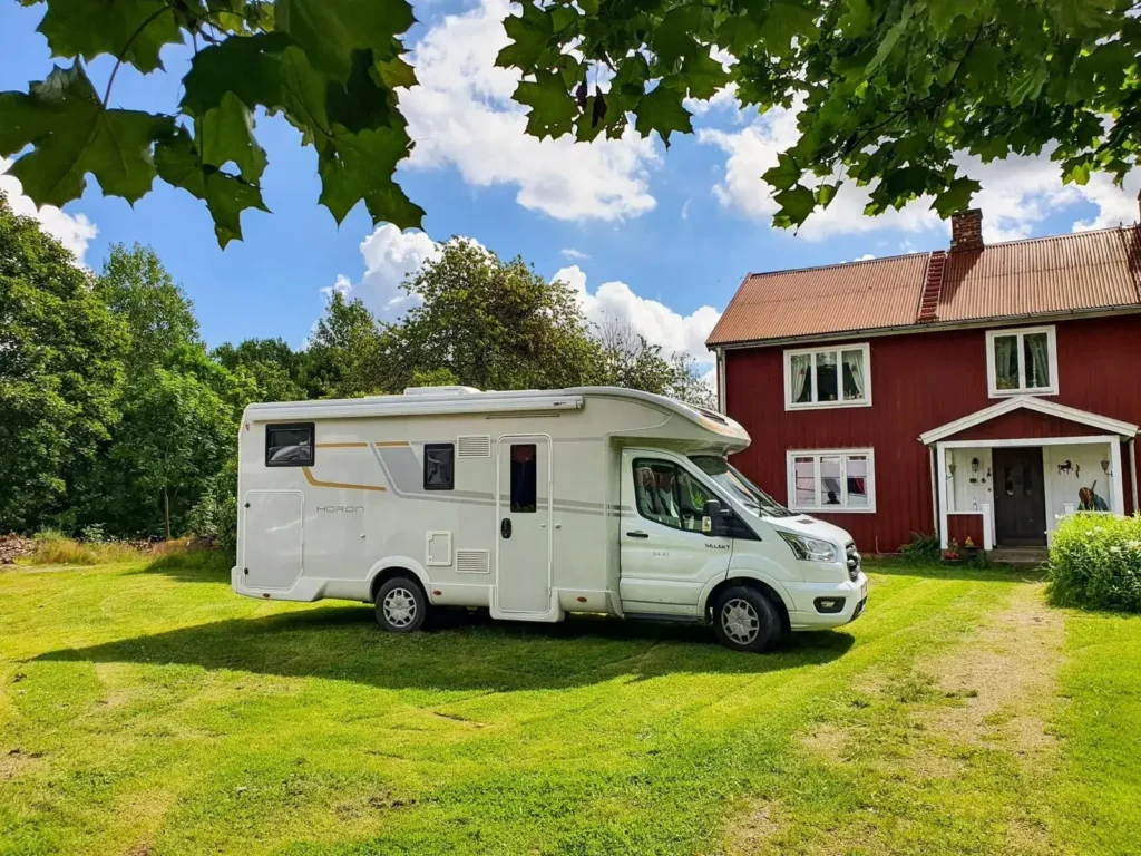 Biały kamper stojący na zielonym trawniku przy czerwonym, drewnianym domu w otoczeniu drzew