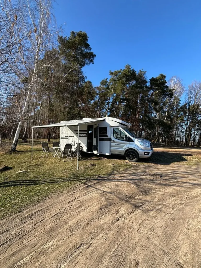 Kamper Roller Team Zefiro z markizą i zestawem wypoczynkowym na postoju w lesie Kamper Roller Team Zefiro z rozłożoną markizą i stolikiem na tle lasu