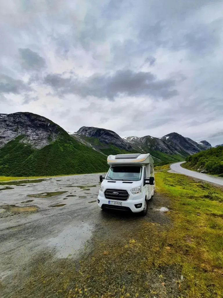Biały kamper zaparkowany na górskiej trasie w deszczowy dzień, otoczony zielonymi zboczami i krętą drogą