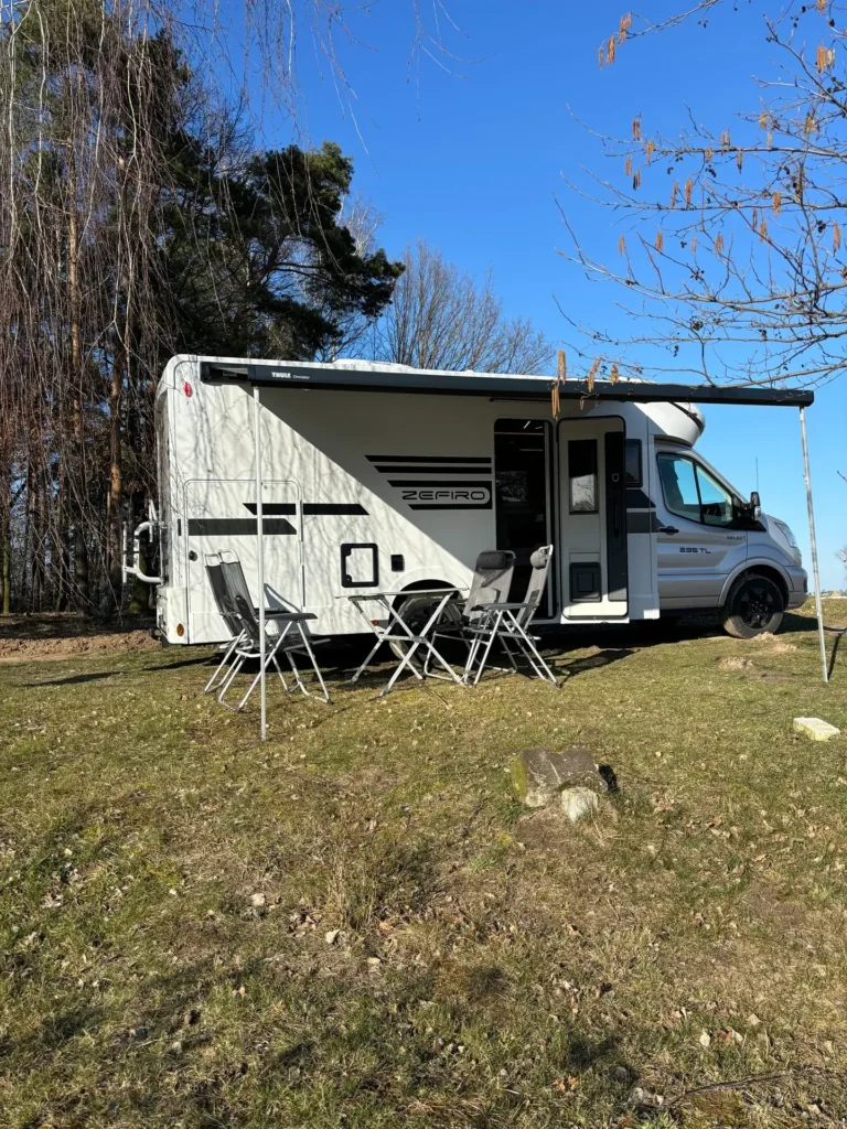 Zefiro 2025 – kamper z zestawem kempingowym gotowy do biwakowania Kamper Roller Team Zefiro 2025 z rozłożoną markizą i krzesłami kempingowymi, ustawiony na trawie pod lasem