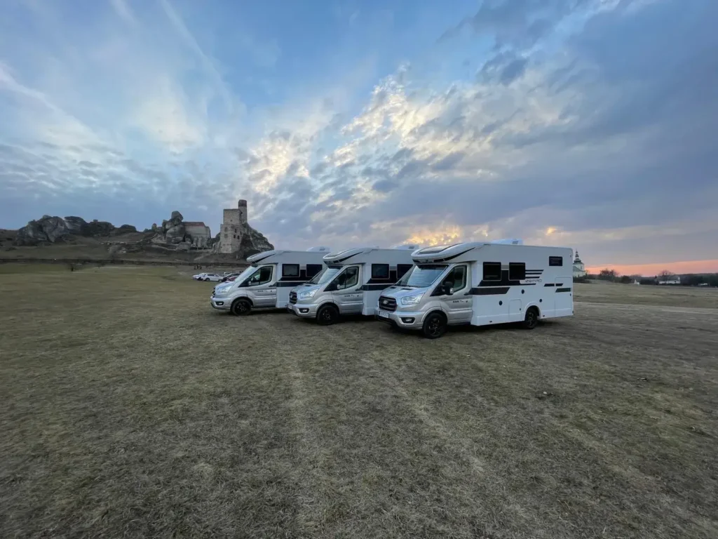 Kampery Roller Team Zefiro na tle zamku – gotowe na podróż Trzy kampery Roller Team Zefiro zaparkowane na otwartej przestrzeni przy zamku o zachodzie słońca